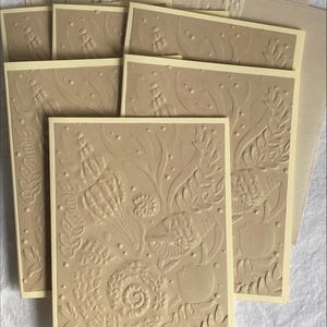 May include: A collection of beige embossed greeting cards with a cream-colored border. The cards feature a detailed underwater design with seashells, seaweed, and bubbles. Several envelopes are also visible.