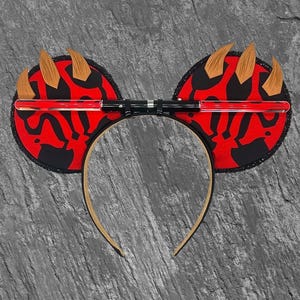 May include: A themed headband with red and black circular ears with a pattern. The ears have brown horns and a red and black lightsaber design. The headband is tan.