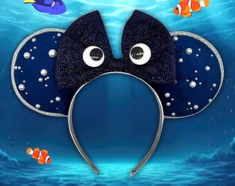 May include: Navy blue mouse ear headband with a sparkly bow and round, white-eyed embellishments. The ears are navy blue with silver trim and pearl accents. The background features cartoon fish and an underwater scene.