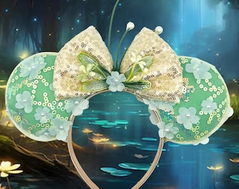 May include: A whimsical headband featuring large, round ears in a light green hue, adorned with delicate floral embellishments and a shimmering gold sequined bow. The headband is accented with small pearls and a gold-toned band.