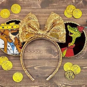 May include: Gold sequined headband with a large bow and character ears featuring Robin Hood and Sir Hiss. The ears are round with gold trim and character illustrations. Gold coins are scattered around the headband on a wooden surface.
