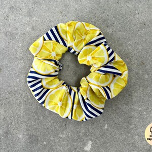 Op de afbeelding: Een gele en blauwe stoffen scrunchie met een citroenprint. De scrunchie is gemaakt van een zachte, rekbare stof en is perfect om haar op zijn plaats te houden.