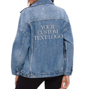 May include: Pale blue denim jacket adorned with scattered rhinestone details. The back of the jacket displays the embroidered text "YOUR CUSTOM TEXT/LOGO". It has a classic design with button fastenings and pockets.