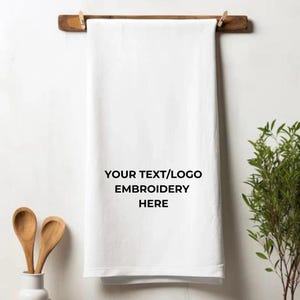 May include: A white kitchen towel with the text "YOUR TEXT/LOGO EMBROIDERY HERE" is hanging from a wooden rod with two wooden clothespins. Two wooden spoons are in a white vase.