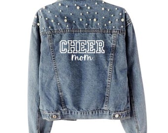 May include: Pale blue denim jacket adorned with faux pearls on the upper back. The words "CHEER mom" are printed in white on the back. The jacket has a classic button-up design with pockets and a collar. The jacket is hung on a hanger.