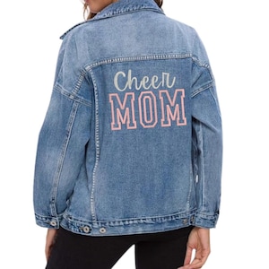 May include: Light blue denim jacket with the words "Cheer MOM" printed on the back in a pink and white design. The jacket has a classic collar, button closures, and pockets.