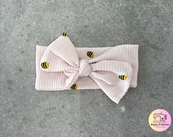 May include: A pink ribbed headband with a bow and a bee pattern. The headband is decorated with small yellow and black bees.