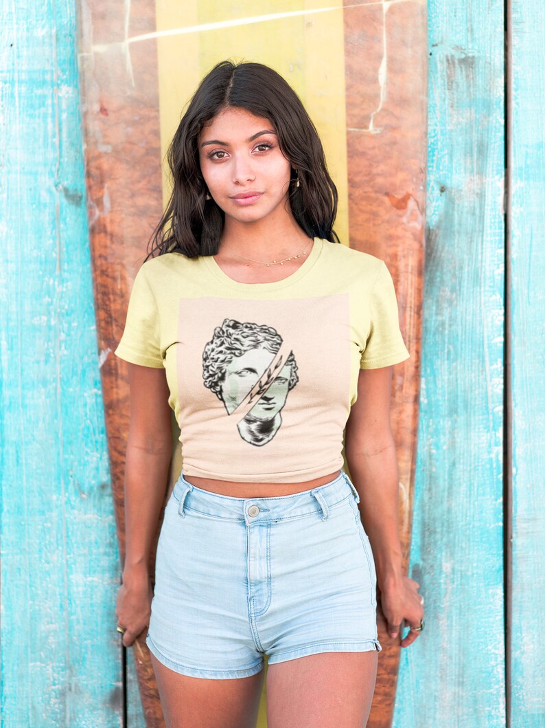 May include: A woman wearing a light yellow t-shirt with a graphic of a broken sculpture of a face. She is wearing light blue denim shorts and standing in front of a colorful wooden wall.