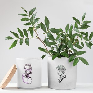 May include: Two white ceramic candle jars with a gray glaze. The jars have a graphic design of a bust of a person with a blindfold. One jar has a bust of a man, the other a bust of a woman. The jars are sitting on a white surface with a green leafy plant in a white ceramic pot.