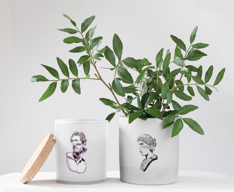May include: Two white ceramic candle jars with a gray glaze. The jars have a graphic design of a bust of a person with a blindfold. One jar has a bust of a man, the other a bust of a woman. The jars are sitting on a white surface with a green leafy plant in a white ceramic pot.