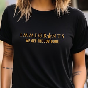 Peut inclure: T-shirt noir avec texte doré « IMMIGRANTS WE GET THE JOB DONE » et une étoile. Le t-shirt a des manches courtes et un col rond.
