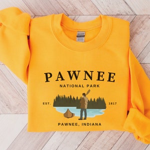 Op de afbeelding: Geel sweatshirt met een grafisch ontwerp van een nationale parkscène. De tekst luidt "Pawnee National Park Est. 1817 Pawnee, Indiana".