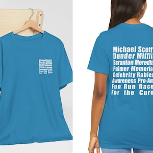 Può includere: Una maglietta blu chiaro con testo bianco che recita "Michael Scott's Dunder Mifflin Scranton Meredith Palmer Memorial Celebrity Rabies Awareness Pro-Am Fun Run Race For the Cure".