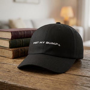 May include: Black baseball cap with the text "FIST MY BUMP" in white. The cap is sitting on a wooden surface next to a stack of books. The background includes a couch and a lamp.