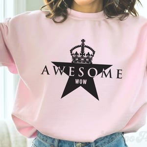 Può includere: Felpa rosa chiaro con grafica a stella nera. La stella ha la scritta "AWESOME" sopra e "WOW" sotto. Una corona nera è sopra la stella. La felpa è in stile casual.