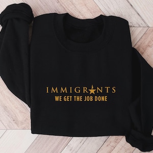 Peut inclure: Sweat-shirt noir à col rond avec les mots "IMMIGRANTS" au-dessus de "WE GET THE JOB DONE" en lettres dorées. Une étoile dorée est entre les mots "IMMIGRANTS" et "WE". Le sweat-shirt est plié sur une surface claire.