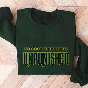 May include: Dark green sweatshirt with the phrase "NO GOOD DEED GOES UNPUNISHED" in yellow and green lettering. The sweatshirt is folded on a light brown wooden surface.