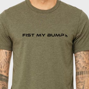 May include: Olive green t-shirt with the text "FIST MY BUMP" in black, with a small graphic. The shirt has short sleeves and a crew neck. The fabric appears to be a soft, heathered material.
