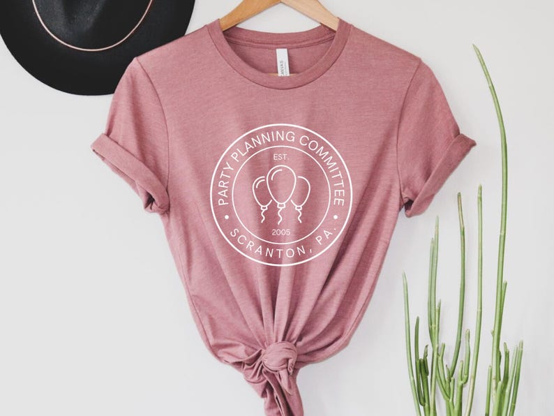May include: A pink t-shirt with a white circle logo that says "Party Planning Committee Est. 2005 Scranton, PA" with a graphic of three balloons.