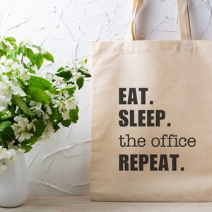 May include: A beige canvas tote bag with black text that reads "EAT. SLEEP. the office REPEAT."