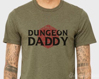 以下が含まれることがあります： オリーブグリーンのTシャツ。中央に赤い幾何学模様があり、黒字で「DUNGEON DADDY」と書かれています。Tシャツは柔らかい素材でできており、半袖です。デザインは胸の中央に配置されています。
