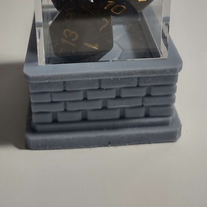 May include: A clear acrylic cube with a black base sits on a gray brick-like platform. Inside the cube are six black dice with gold numbers. The dice show the numbers 3, 5, 6, 7, 10, and 13.
