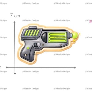May include: A gray and black toy ray gun with green and yellow accents, measuring 10 cm long and 7 cm high. The ray gun has a light brown outline and the text "MDC057".