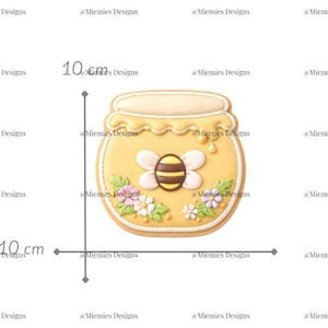 Puede incluir: Una galleta con forma de tarro de miel, de aproximadamente 10 cm de alto y ancho, decorada con glaseado amarillo y una abeja. La galleta está adornada con flores blancas y rosas y el texto "MDC 151".