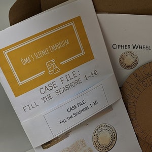 May include: A brown wooden cipher wheel with a white alphabet printed on it. The wheel is in a white box with other items, including a white envelope with the text "Shoe and Seashore Envelopes with Encrypted Labels" printed on it.
