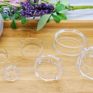 May include: Clear acrylic ring-shaped display stands of varying sizes are arranged on a wooden tray. The stands are designed to hold and showcase small items. A decorative pot with a plant and flowers are in the background.