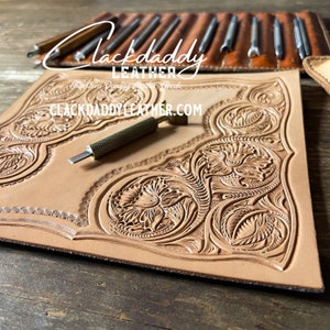 May include: A close-up of a piece of leather with an intricate floral design carved into it. The leather is light brown and the design is a light tan color. A small metal tool is lying on the leather.