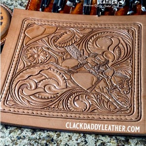 Op de afbeelding: Een gedetailleerd, met de hand bewerkt leren stuk met een cowboy op een paard, omringd door bloemen en krulontwerpen. Het bruine leer heeft een gesneden textuur met de tekst "Clackdaddy Leather" en "ClackdaddyLeather.com".