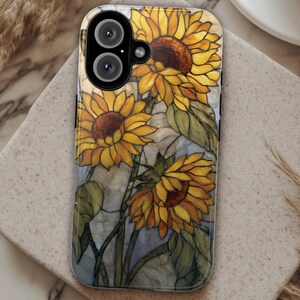 May include: A phone case with a stained-glass effect sunflower design. The case displays yellow sunflowers with brown centres and green leaves on a light blue and grey backdrop. The case has a glossy finish and a camera cutout.