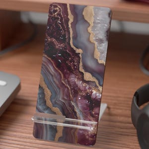 May include: A rectangular phone stand with a marbled design in shades of burgundy, gray, and gold. The stand is made of a hard material and is propped up by a clear plastic stand. The stand is designed to hold a phone or tablet.
