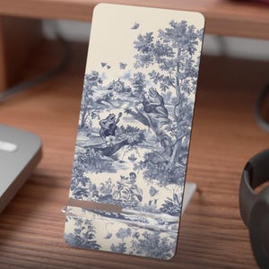 Può includere: Un supporto piatto stampato per display mobile con sfondo crema e un design toile de Jouy blu e bianco con rane che suonano strumenti musicali. Il supporto è su una scrivania di legno. Il testo "Flat Printed Mobile Display Stand" è in basso.