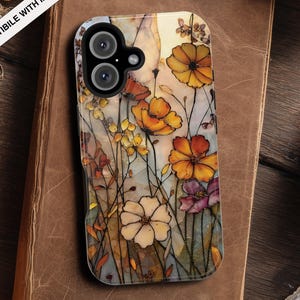 Puede incluir: Una funda de teléfono floral con un diseño de acuarela con flores naranjas, amarillas y moradas. La funda es compatible con MagSafe y descansa sobre un libro antiguo. Tiene un recorte para una cámara de doble lente.