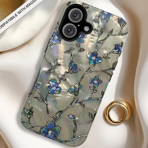 May include: A phone case with a pearlescent, iridescent finish featuring a dark-outlined floral vine pattern with blue petals and small white centers. The case has a black camera cutout and includes the text COMPATIBLE WITH MAGSAFE.