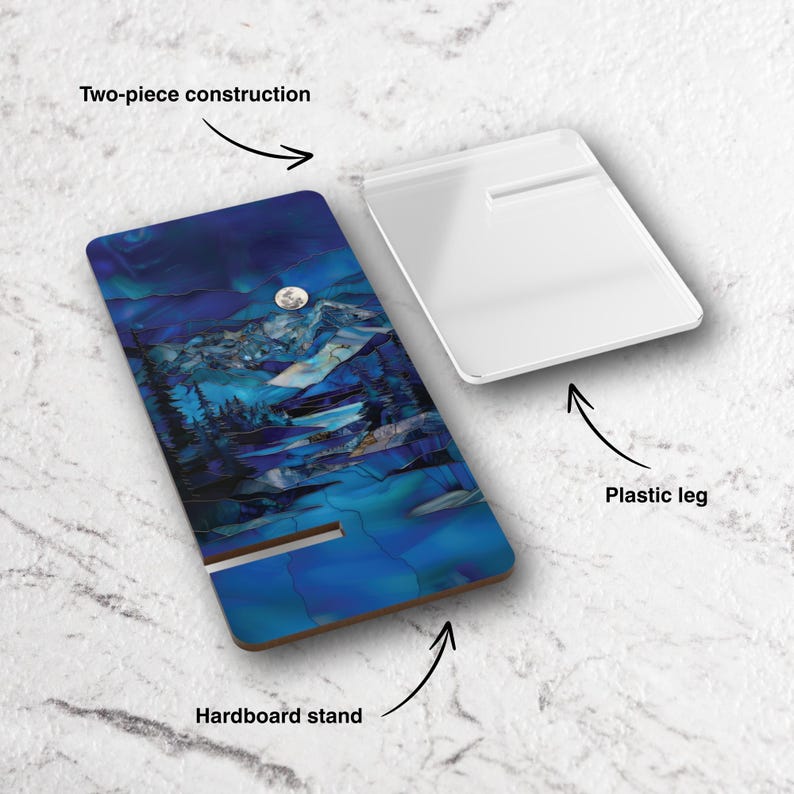 May include: A two-piece hardboard phone stand with a landscape design. The stand features a vibrant blue and teal mountain scene with a full moon. A separate white plastic leg provides support. The stand is designed to hold a phone.