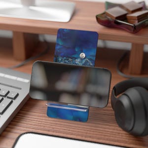 May include: A smartphone rests on a blue and white patterned phone stand. The stand has a matching card holder with a silver moon accent. The scene is set on a wooden desk with a keyboard and headphones.