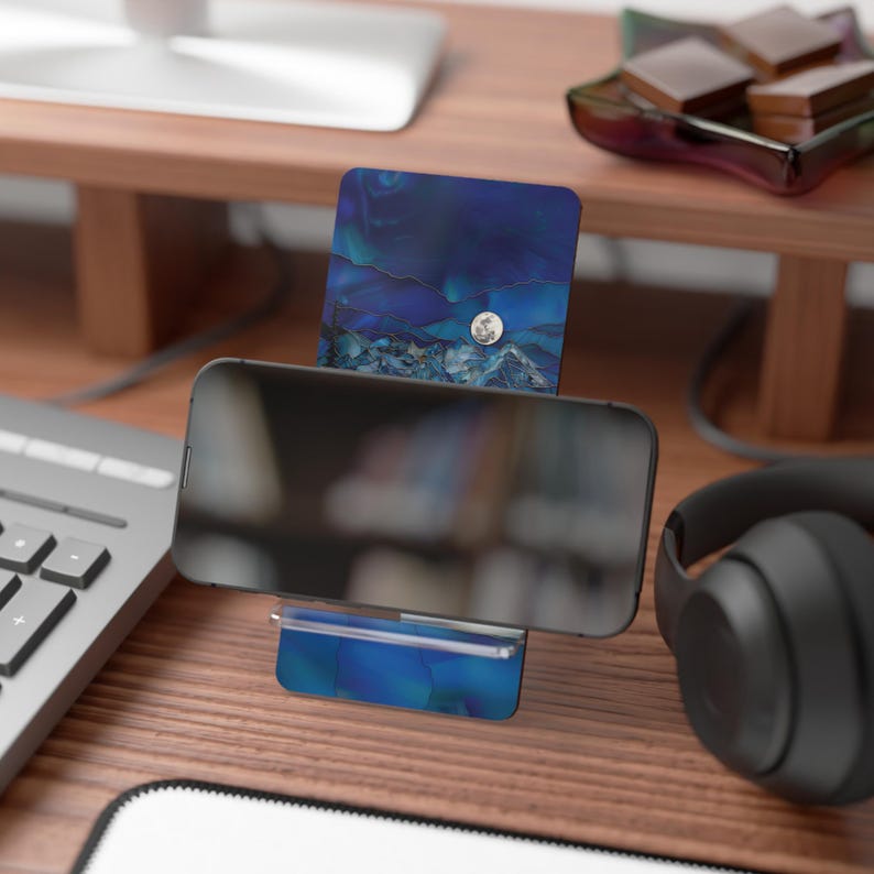May include: A smartphone rests on a blue and white patterned phone stand. The stand has a matching card holder with a silver moon accent. The scene is set on a wooden desk with a keyboard and headphones.
