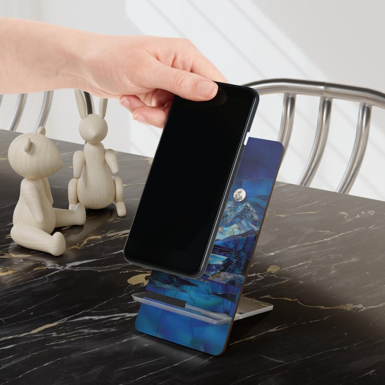 May include: A black smartphone is placed on a blue and white phone stand with a mountain and moon design. The stand has a clear plastic shelf. Two wooden figurines of a bear and a rabbit are on the table.
