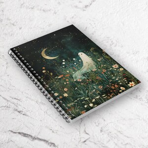 May include: Spiral-bound notebook featuring a whimsical illustration. The cover depicts a serene night scene with a crescent moon, stars, and a ghostly figure in a field of wildflowers. The color palette includes dark blues, greens, and touches of gold.