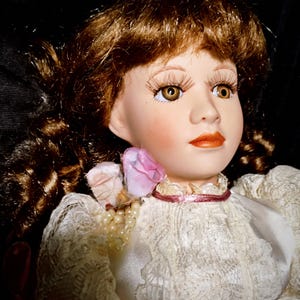 May include: A porcelain doll with long brown hair, wearing a white lace dress with a pink flower accent. The doll has a painted face with brown eyes and a pink mouth.
