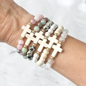 May include: A collection of beaded bracelets featuring cream-colored cross charms and various gemstone beads in shades of pink, green, white, and speckled black. Gold-colored spacers add detail to the bracelets.