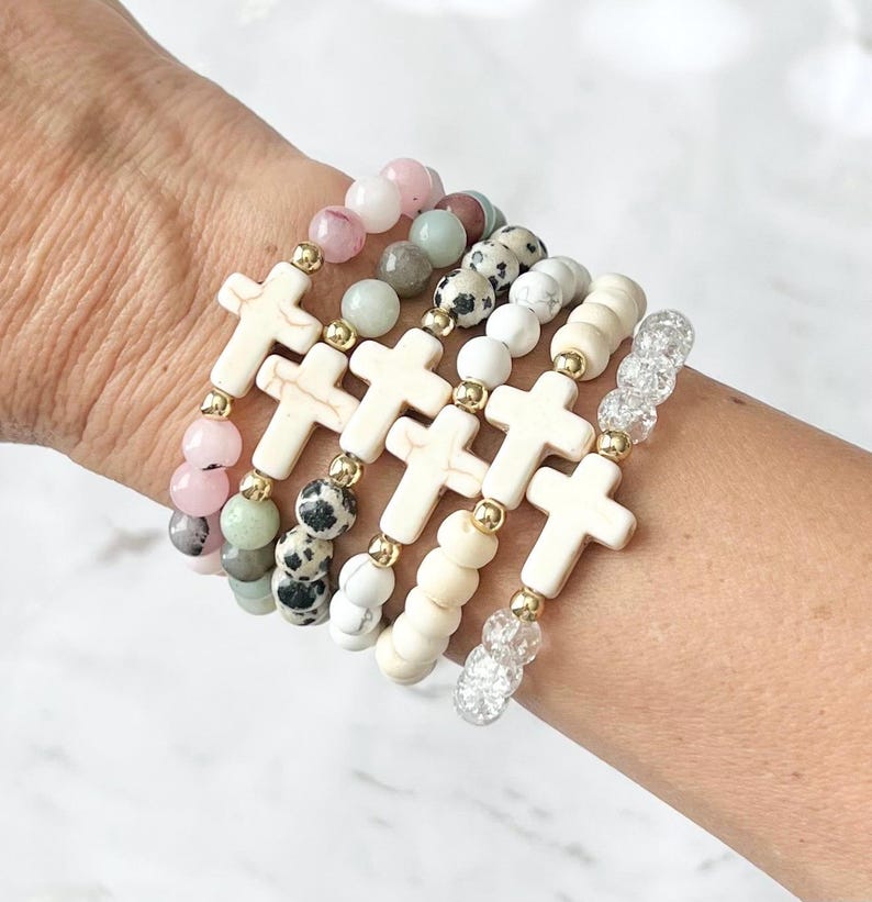 May include: A collection of beaded bracelets featuring cream-colored cross charms and various gemstone beads in shades of pink, green, white, and speckled black. Gold-colored spacers add detail to the bracelets.