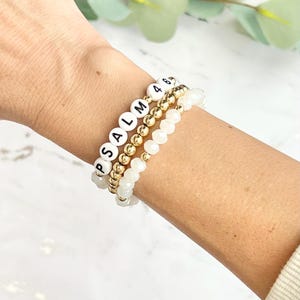 May include: Three bracelets stacked on a wrist. The top bracelet is made of gold beads with white letter beads spelling "PSALM". The middle bracelet is made of gold beads. The bottom bracelet is made of white beads.