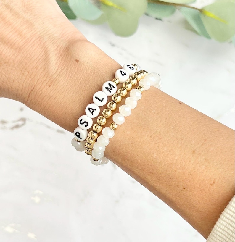 May include: Three bracelets stacked on a wrist. The top bracelet is made of gold beads with white letter beads spelling "PSALM". The middle bracelet is made of gold beads. The bottom bracelet is made of white beads.