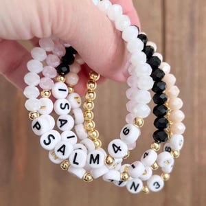 May include: Set of four bracelets with white beads and black lettering. The bracelets spell out "PSALM 4" and "ISAIAH 43".