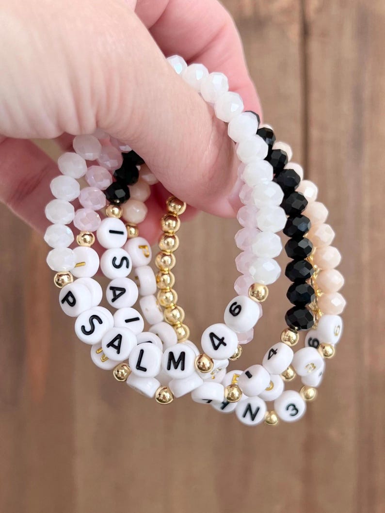 May include: Set of four bracelets with white beads and black lettering. The bracelets spell out "PSALM 4" and "ISAIAH 43".
