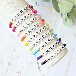 May include: A set of 10 colorful bracelets with white beads spelling out different names, each with a gold bead and a white bead with a paw print. The bracelets are arranged in a row on a white surface.